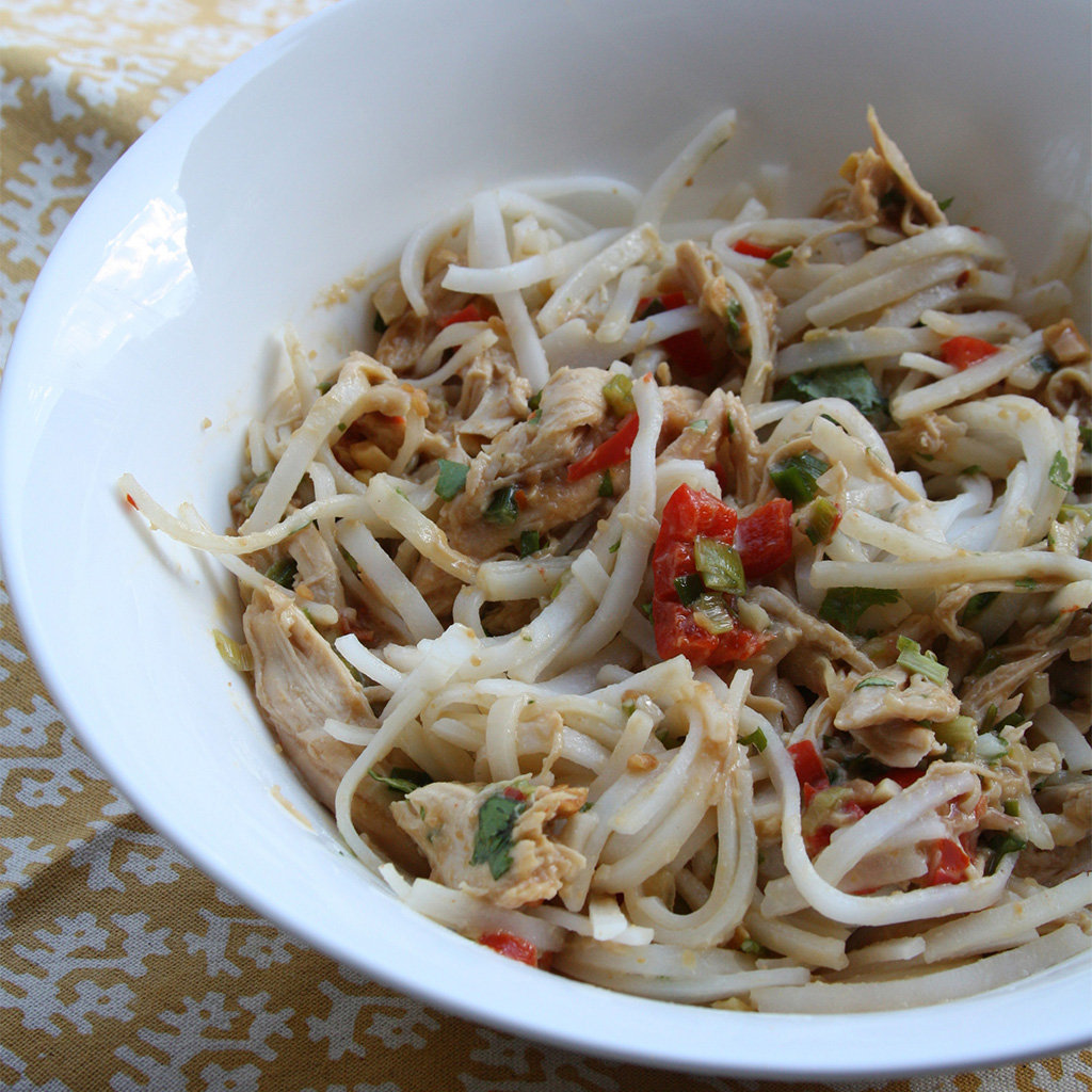 Rice noodles with shredded chicken, bean sprouts and red pepper in peanut sauce in a white bowl.