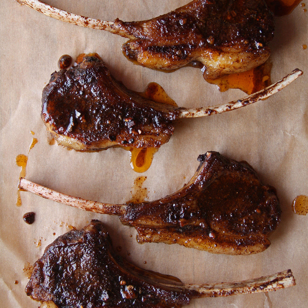 Four coffee-rubbed lamb chops with glistening caramelized crusts on parchment, drizzled with chili oil, shot from above.