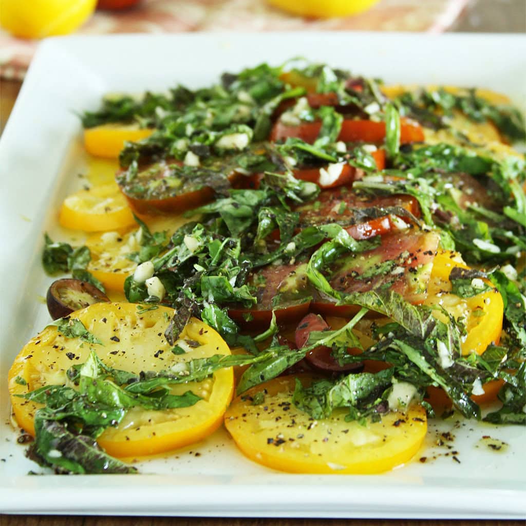 Heirloom yellow and red tomato salad layered with basil and herbed dressing on a square white plate.