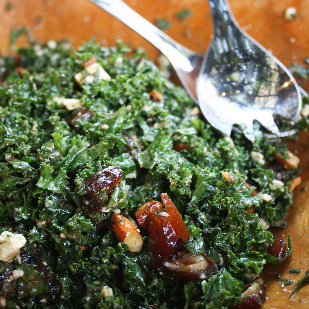 Finely chopped kale salad with sliced dates and almonds being tossed with serving spoons.
