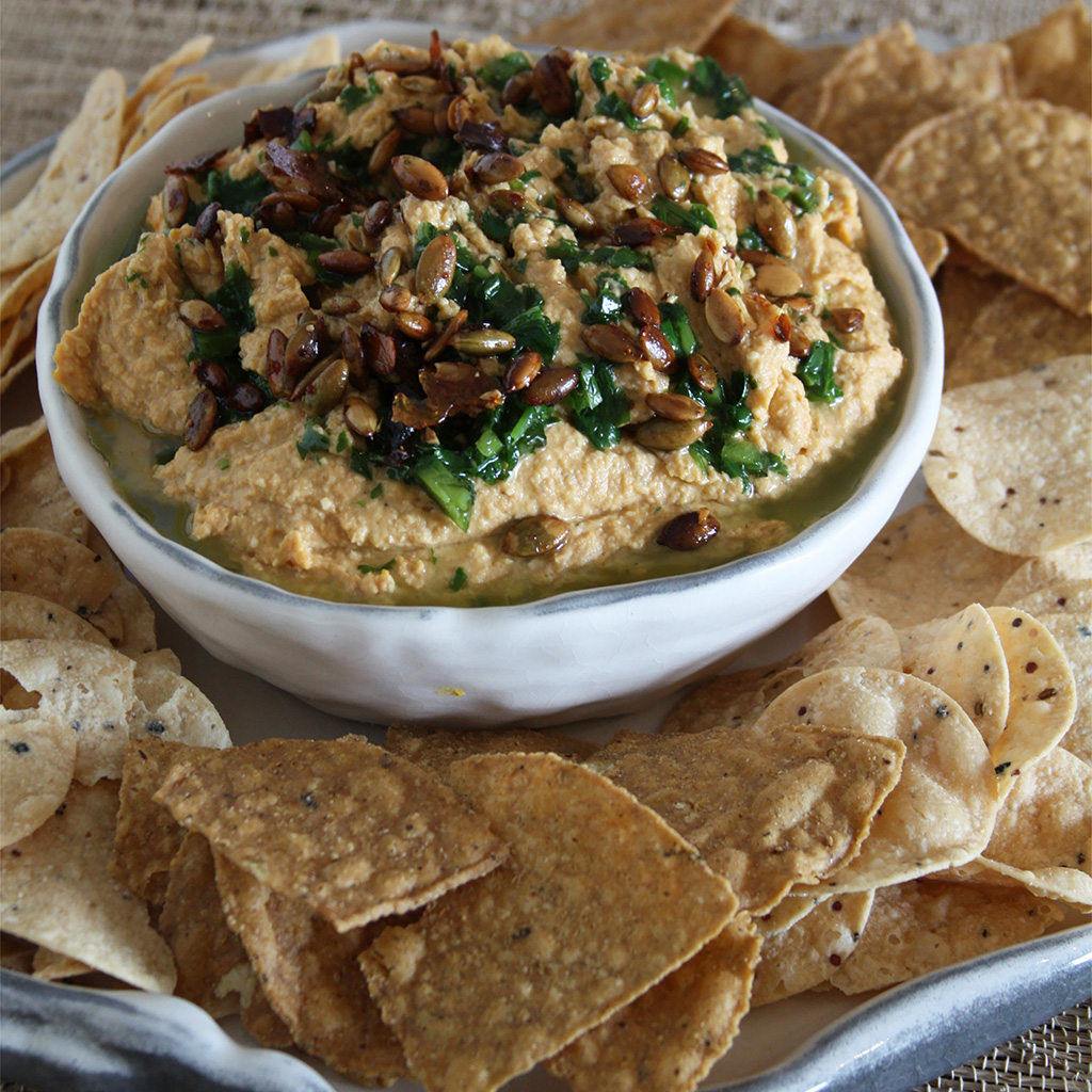 Bowl of pumpkin hummus topped with candied pumpkin seeds and jalapeno cilantro oil, tortilla chips around.