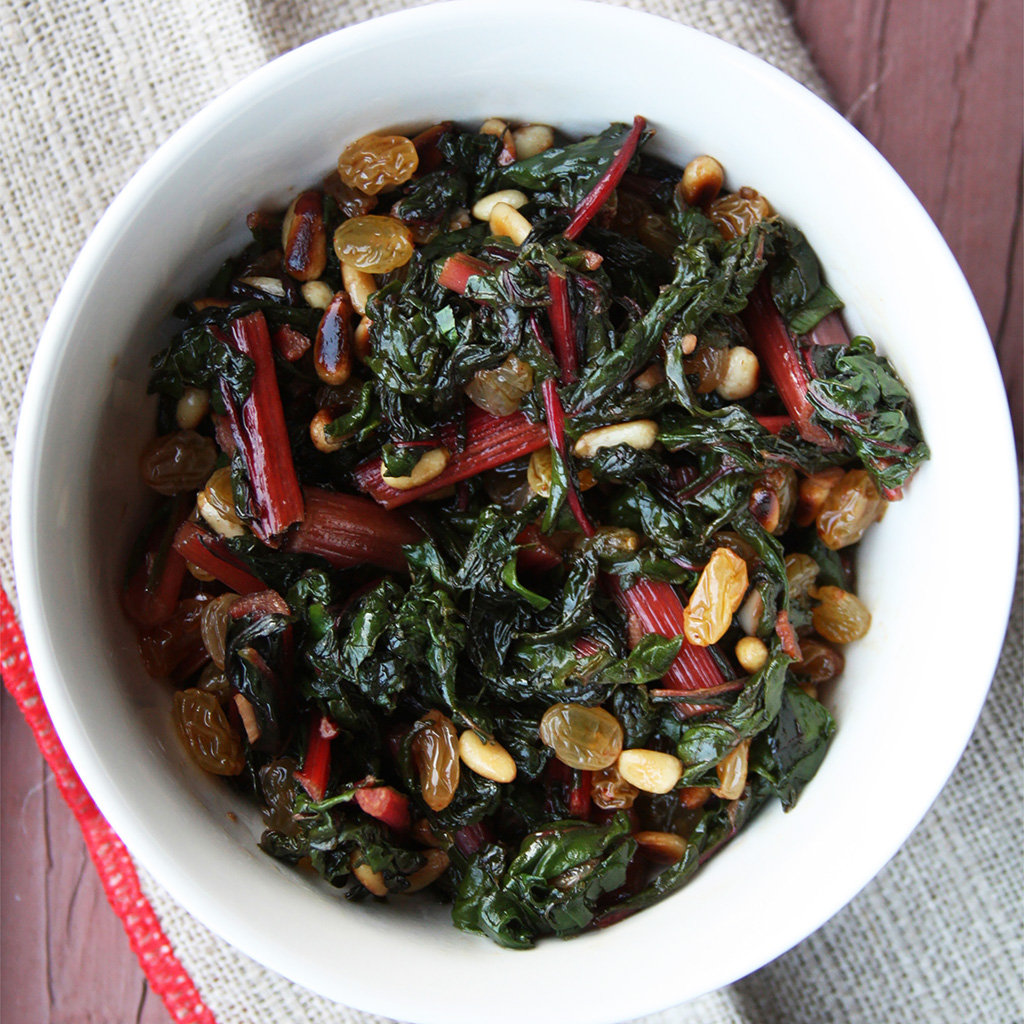 Sauteed rainbow chard with red stems, golden raisins and toasted pine nuts in a white bowl.