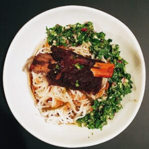 Braised Asian spiced short rib on rice noodles topped with bright green serrano chimichurri in a white bowl.