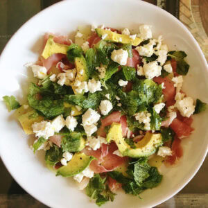 Melon, prosciutto and avocado salad with torn mint and crumbled feta in a white bowl.