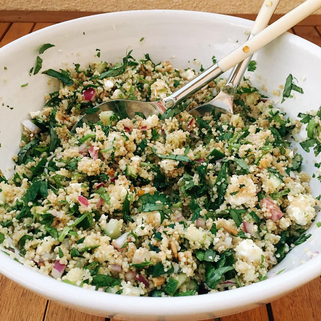 Summer French couscous with parsley, mint, cucumber and feta being tossed in a large white bowl.