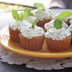Almond cupcakes topped with herb-flecked lemon mint icing and fresh mint leaves on a yellow plate.