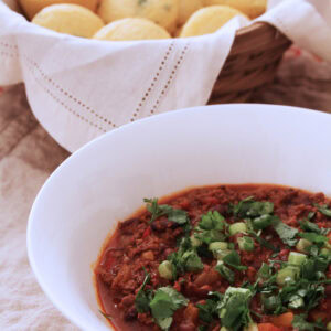 Bowl of black bean chili topped with cilantro and sliced scallions, with cornbread muffins behind.
