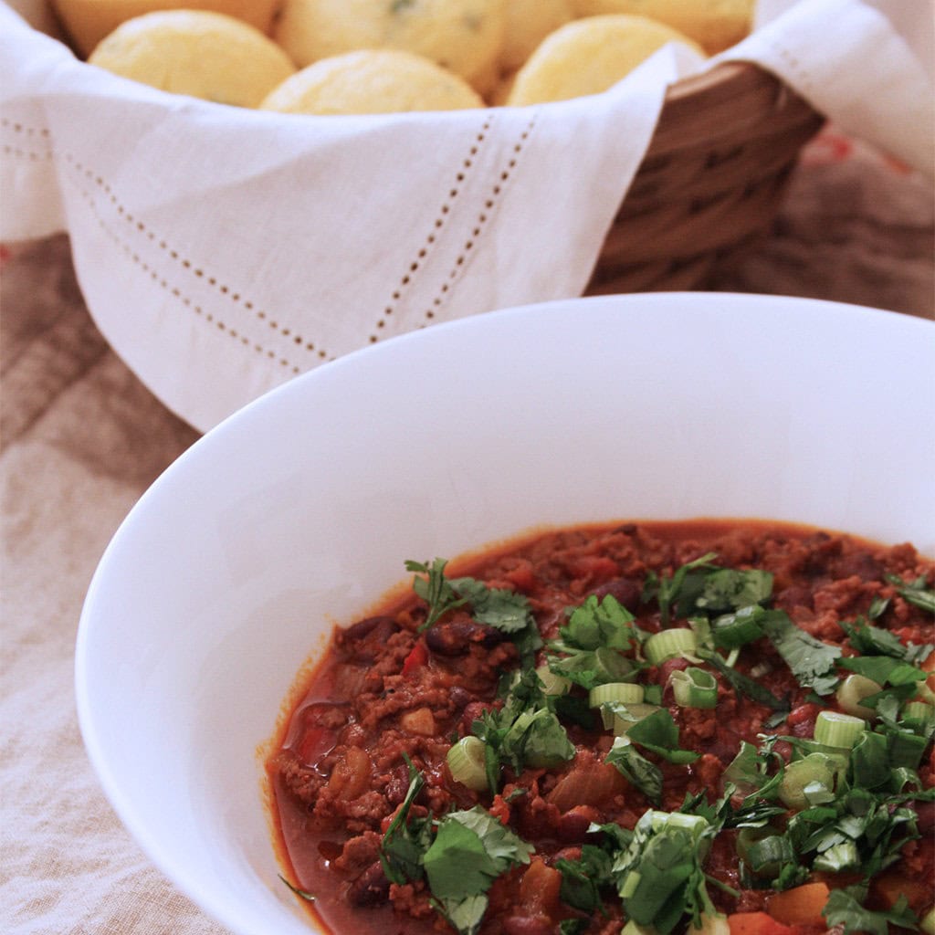 Bowl of black bean chili topped with cilantro and sliced scallions, with cornbread muffins behind.