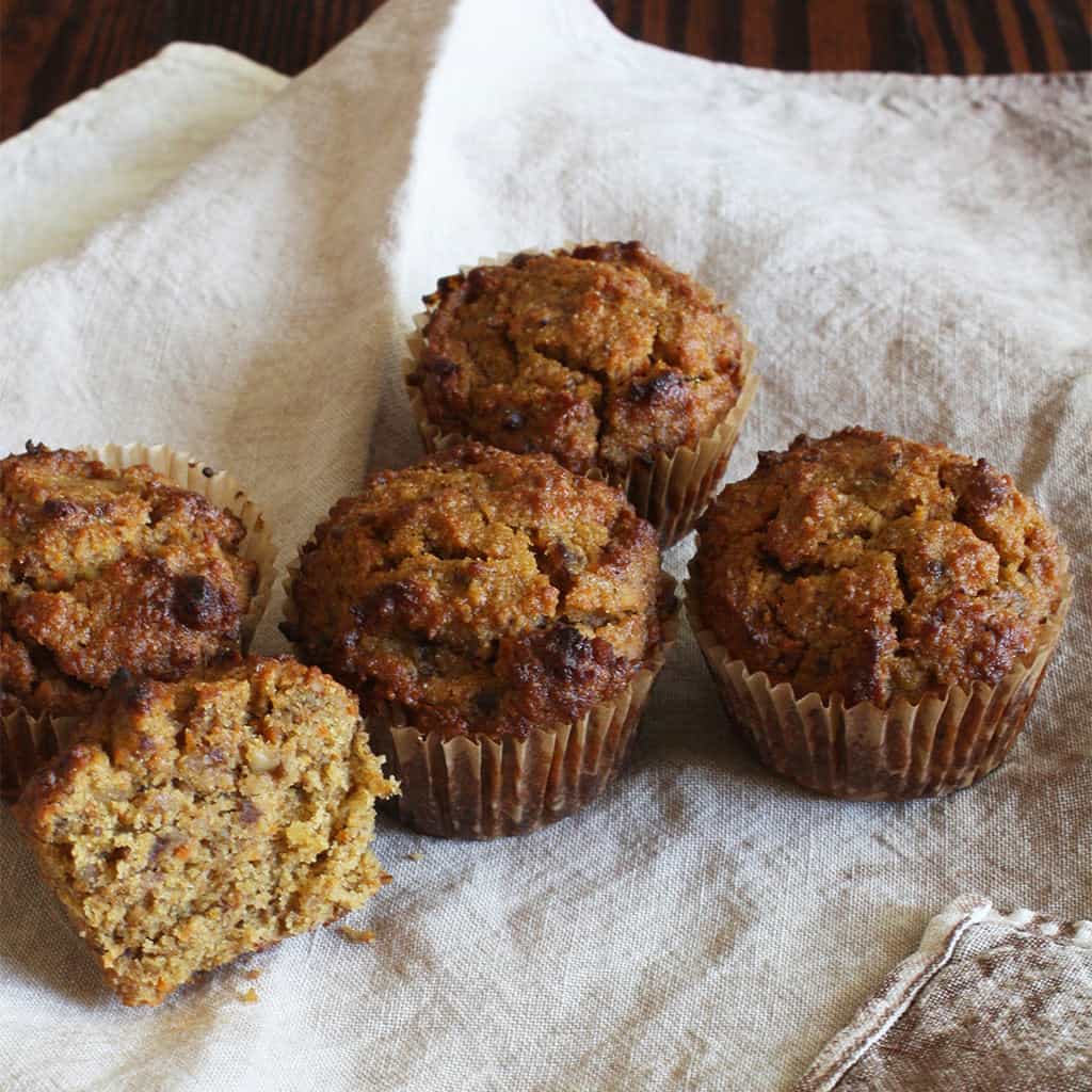 Four carrot and date muffins on a linen cloth, one split open to show the moist crumb.