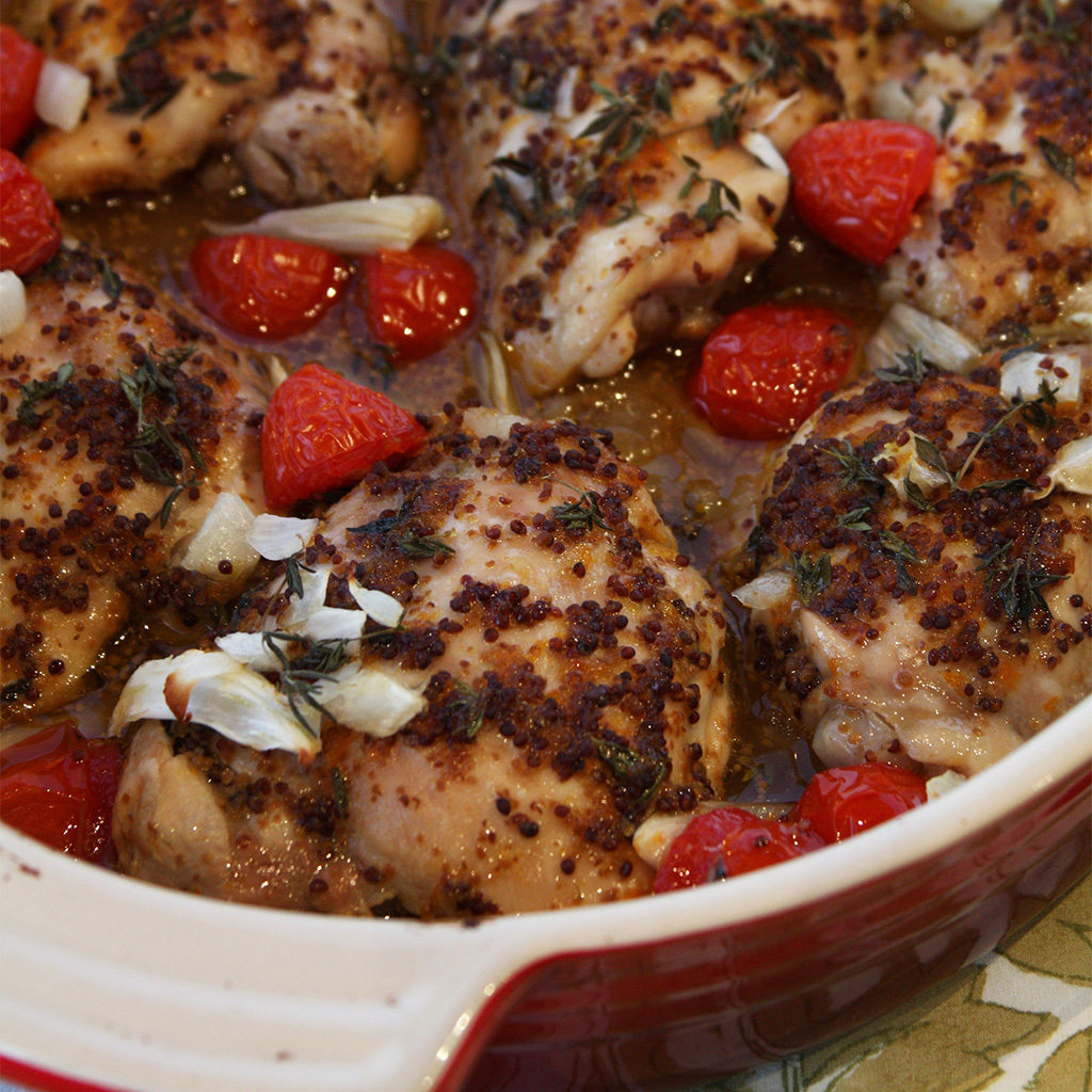 Mustard-marinated chicken thighs baked with blistered cherry tomatoes, garlic and thyme in a red dish.