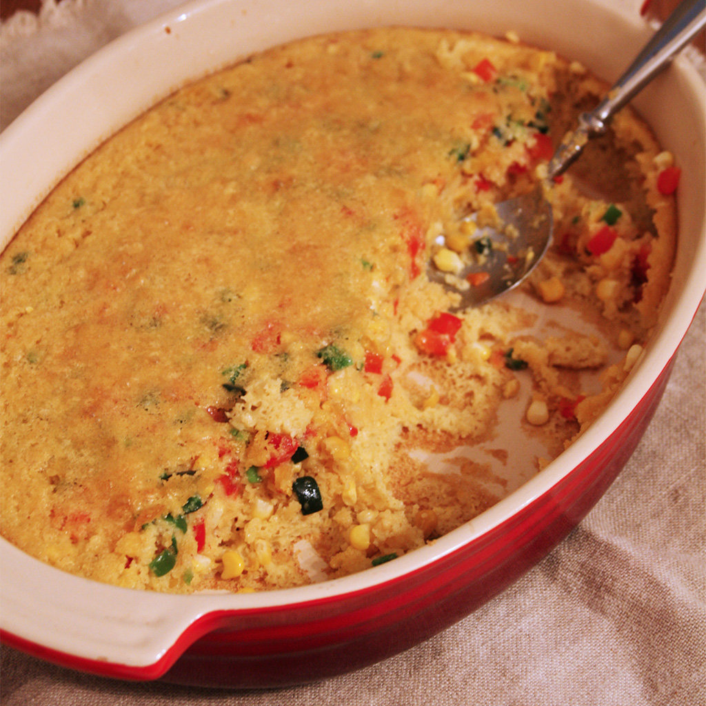 Baked corn pudding flecked with red and green peppers in a red casserole dish with a serving spoon.