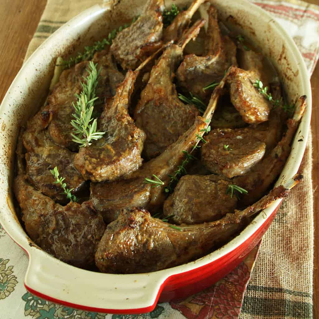 Lamb chops braised with rosemary and thyme in a red and cream oval baking dish.