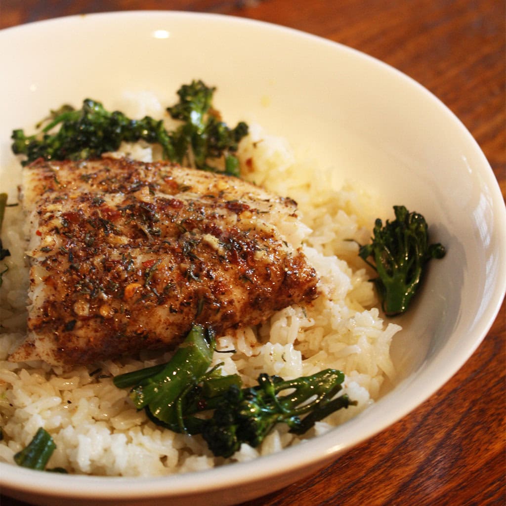 Spice-crusted roasted seabass fillet on herbed rice with charred broccolini in a white bowl.