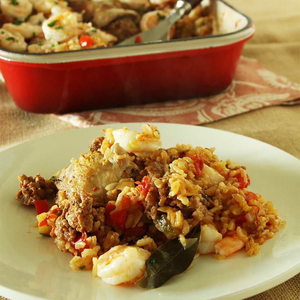 Plated paella with chicken, spicy Italian sausage, shrimp and a bay leaf, red pan behind.