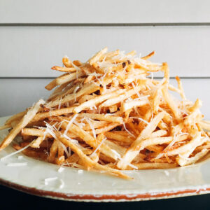 Tall pile of shoestring pomme frites showered with shaved parmesan on a rustic plate.