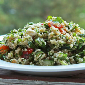 Quinoa tabbouleh with parsley, mint, cucumber and cherry tomatoes heaped in a white bowl outdoors.