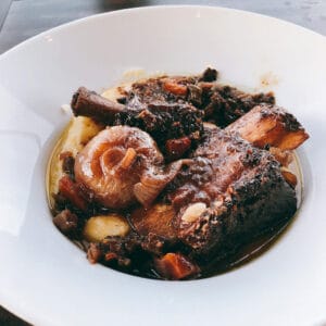 Slow braised short ribs with whole cipollini onions and pan sauce over polenta in a white bowl.