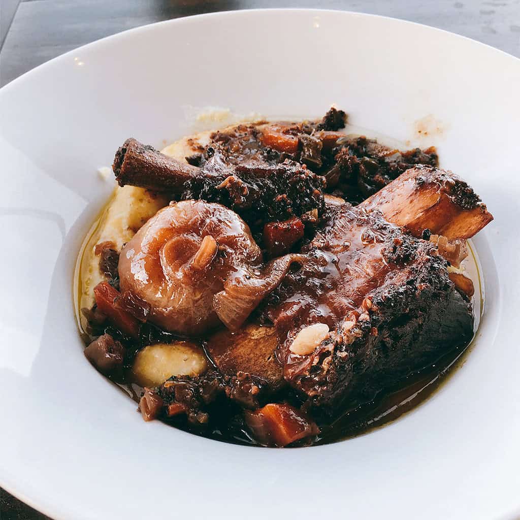 Slow braised short ribs with whole cipollini onions and pan sauce over polenta in a white bowl.