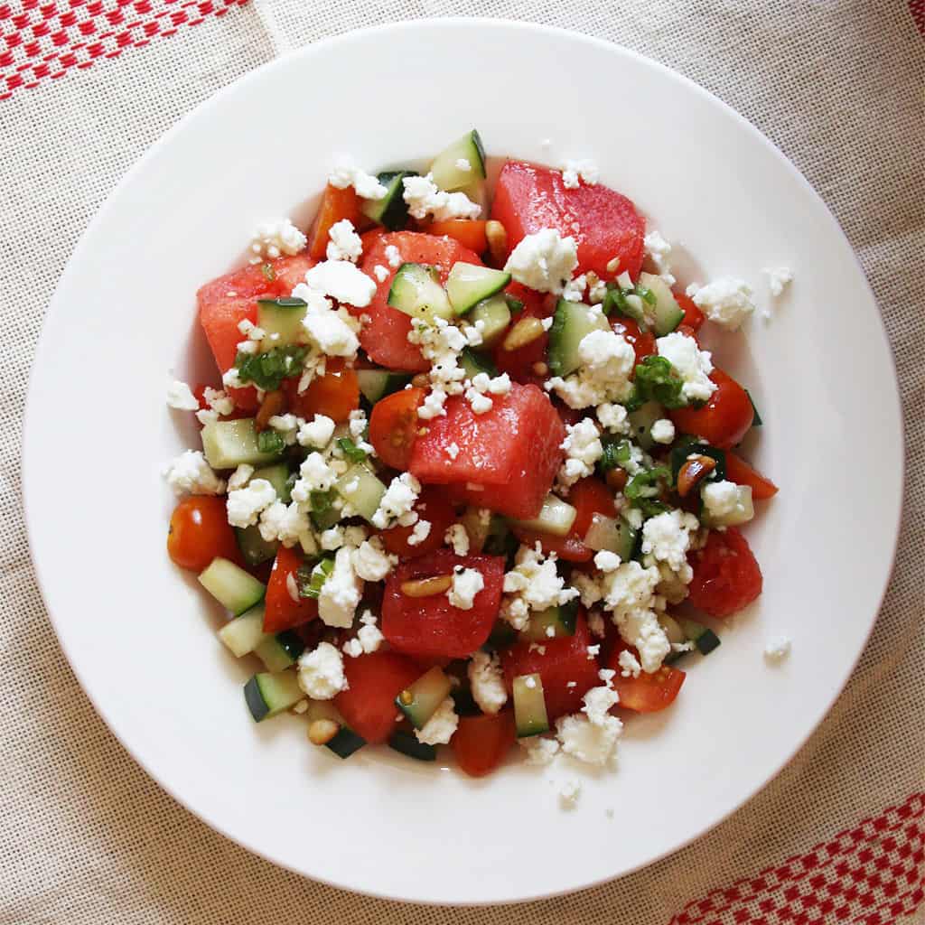 Watermelon salad with cucumber, cherry tomatoes and crumbled feta on a white plate over red and white napkin.