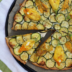 Zucchini and goat cheese tart topped with stuffed squash blossoms, one slice cut, on parchment.