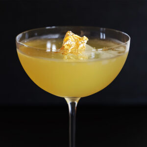 Goldenrod cocktail in a coupe class with gold-leaf garnish against a black background.