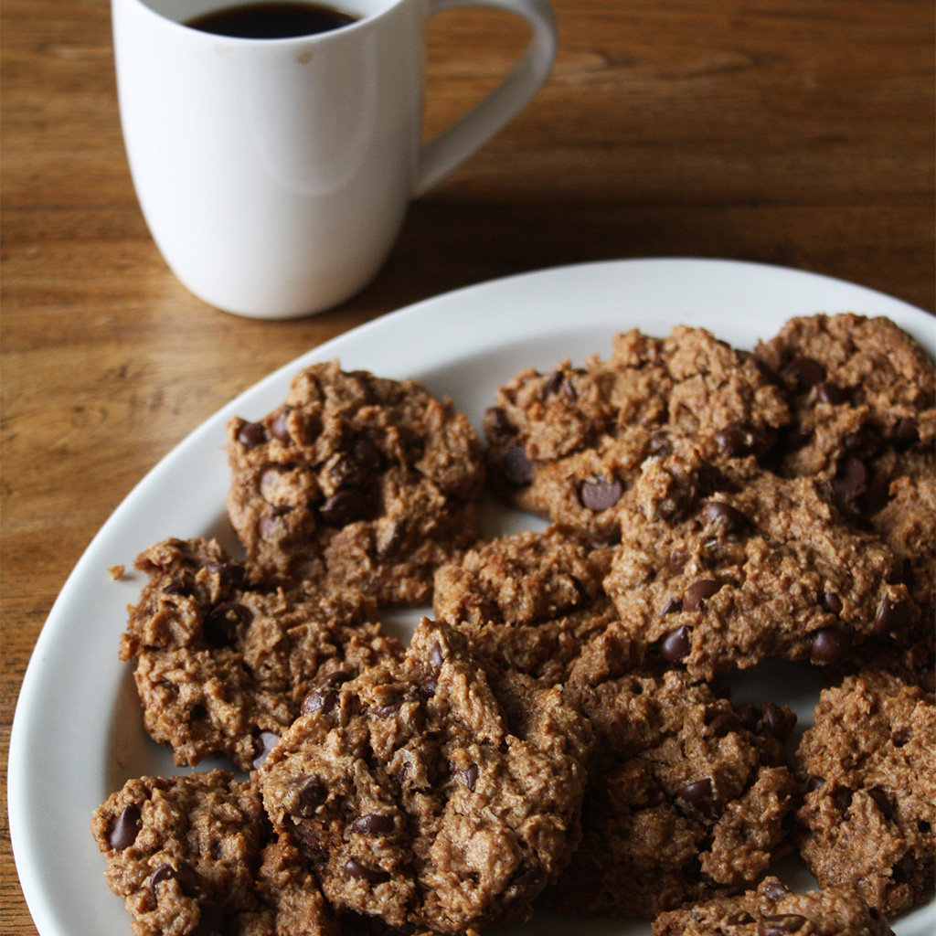 Almond coconut chocolate chip cookies on a white plate beside a mug of black coffee on a wood table.
