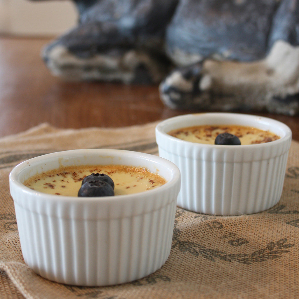 Two ramekins of baked custard topped with a blueberry on a burlap runner.
