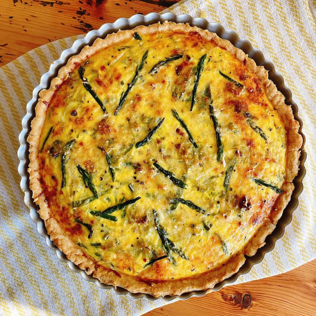 An asparagus, Italian sausage, and green garlic quiche in a fluted tart pan sitting on a yellow striped napkin on a wooden table, shot from above.