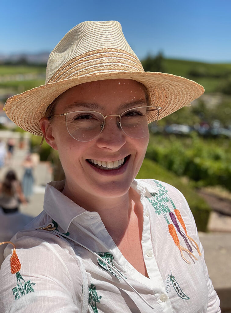 Portrait of Erin wearing a straw hat and a white blouse embroidered with veggies.