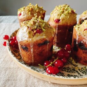 Close-up of red currant and pistachio friands with glaze dripping down their golden sides on a ceramic plate, crushed pistachios on top and red currants beside.