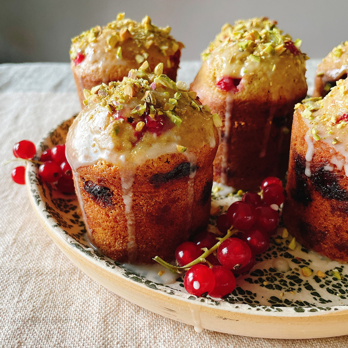 Red currant and pistachio friands with a drippy glaze on a green and white ceramic plate, shot from the side.