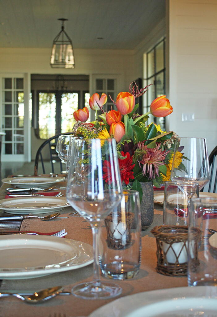 A vase of orange tulips and other flowers on a table set for dinner, surrounded by wine glasses.