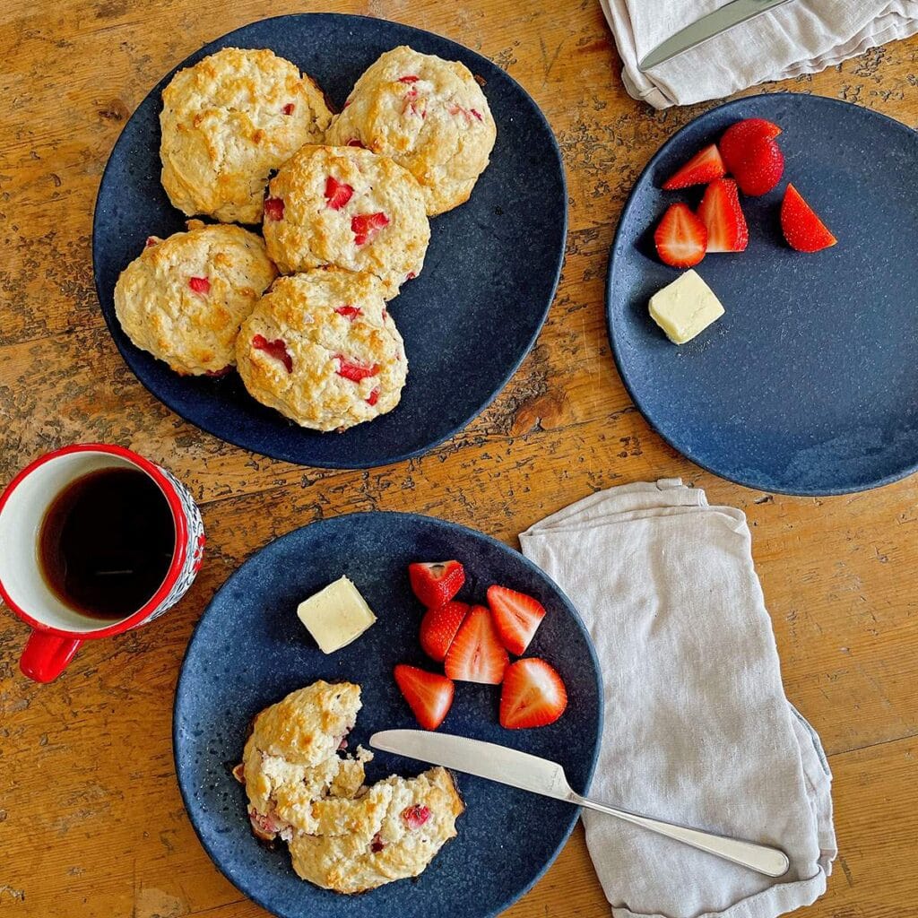 Keto strawberry almond drop biscuits on a black plate, one split on a second plate with butter, strawberries and coffee.